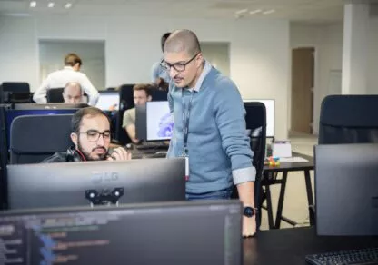 Two members of the Defants team collaborate together: one sits, focused on his laptop, while the other stands beside him, offering support and supervision. The image illustrates dynamic collaboration and team spirit in the field of cybersecurity.