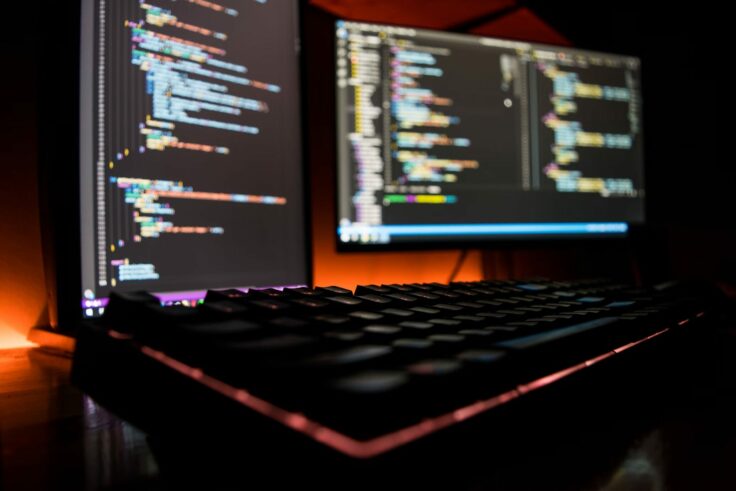 Two bright computer screens display lines of code in a dark atmosphere.