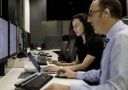 Two people are sitting in front of their computers, each immersed in their work with a serious expression. The office environment around them is calm and professional, emphasizing their concentration and commitment to their tasks.