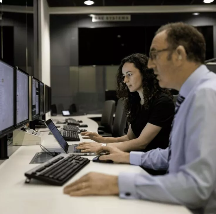 Two people are sitting in front of their computers, each immersed in their work with a serious expression. The office environment around them is calm and professional, emphasizing their concentration and commitment to their tasks.