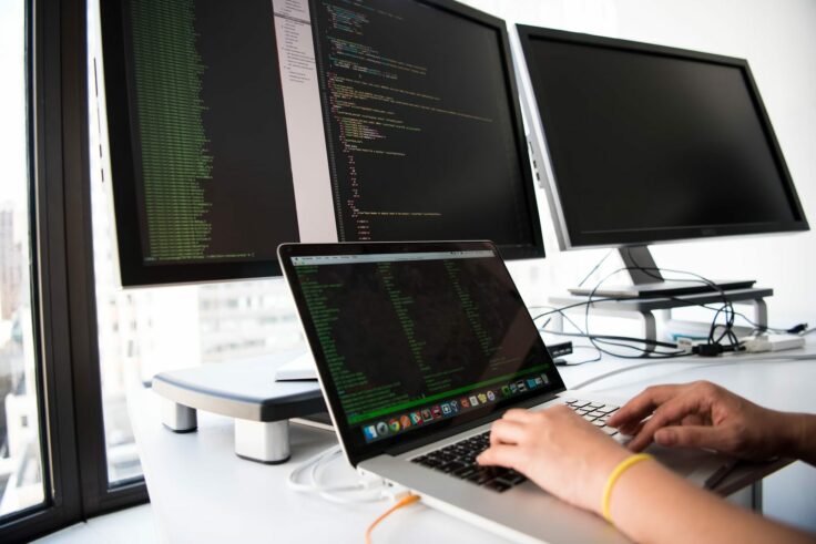 A person's hands are visible in front of several computer screens, each displaying lines of code. The image highlights the intense and important activity of cybersecurity.