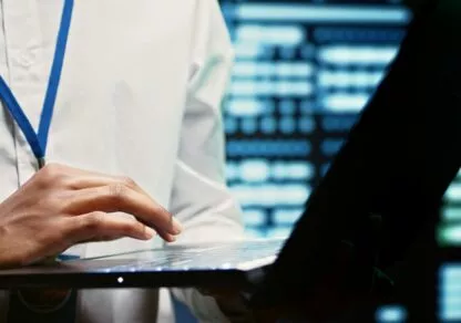 A person stands in front of a row of servers, working on his laptop. The technological environment, with flashing lights from the servers, evokes a professional and dynamic atmosphere.