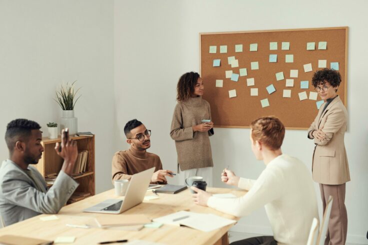 A group of people are gathered around a table, dynamically exchanging ideas. A board is visible in the background, filled with colorful post-its, representing everyone's thoughts and contributions.