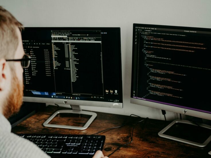 Une personne est assise devant deux écrans d'ordinateurs, concentrée sur son travail.