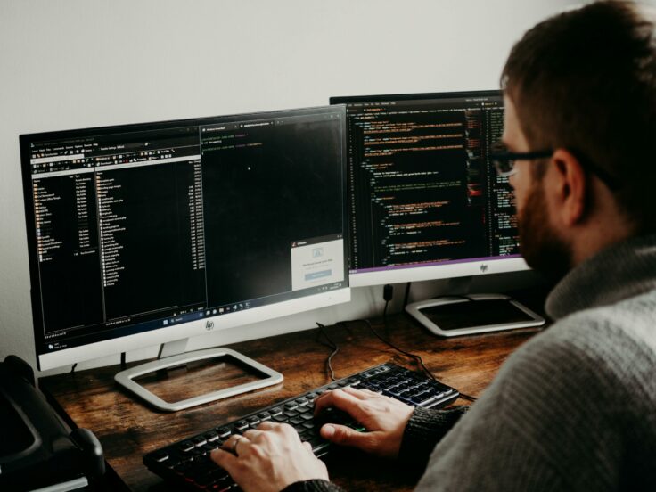 Une personne est assise devant deux écrans d’ordinateur, concentrée sur son travail.