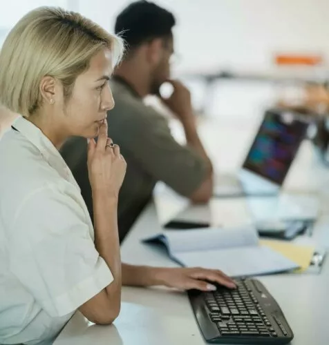 A woman sits at her computer, pensive, thinking about work. Next to her, her colleague is concentrating on his own work.